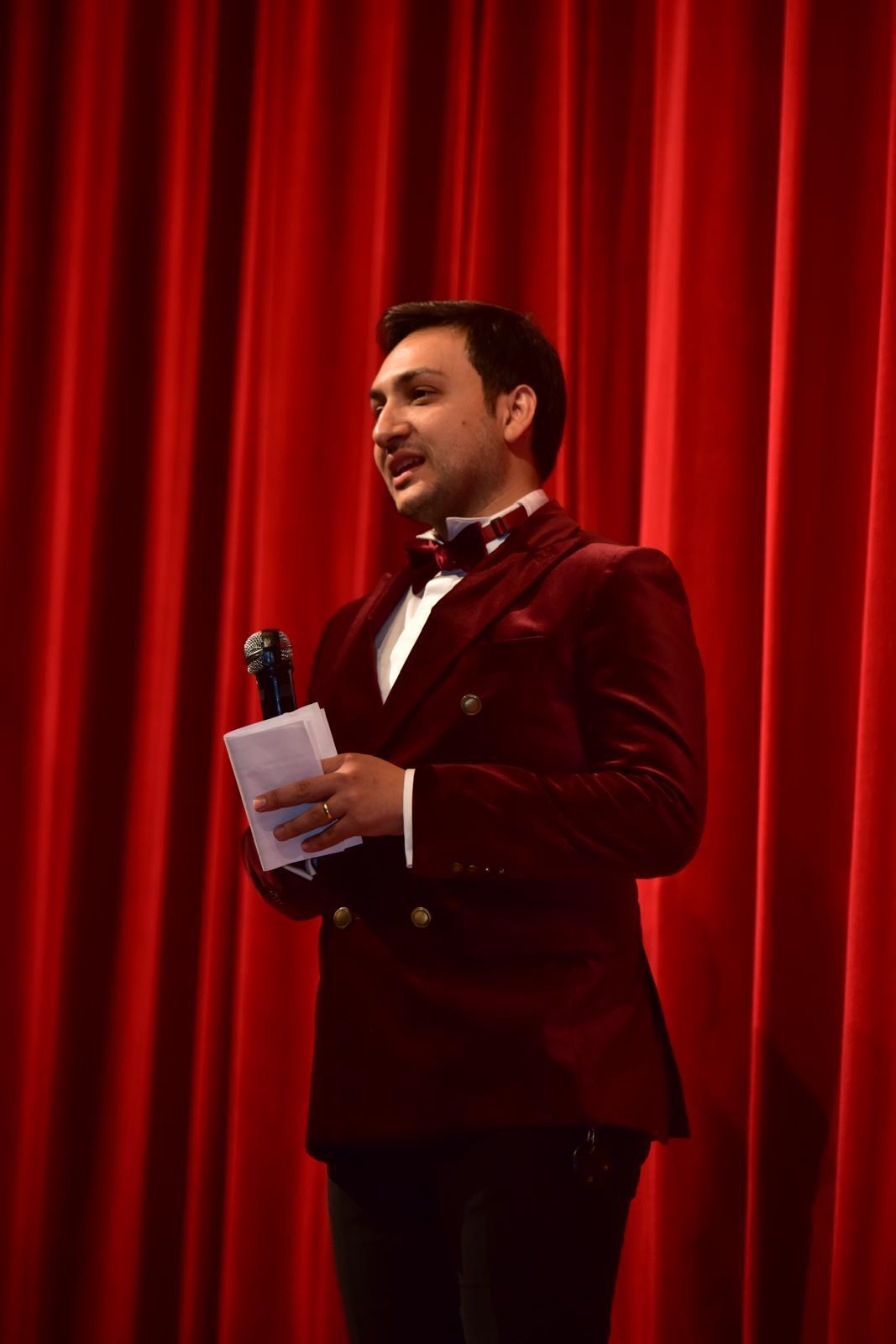 Kırmızı sahne perdesi önünde mikrofon ve sunum notlarıyla sunucu